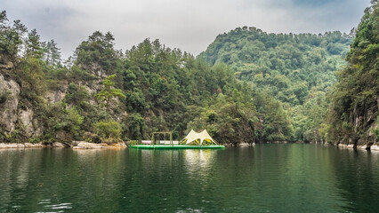 Fototapeta premium The beautiful emerald lake is surrounded by mountains. There is a floating recreation platform with an awning and swings on the water. Thickets of green forest on the mountain slopes.Clouds in the sky
