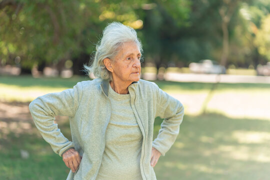 A senior woman with her hands on her hips stands in a park, her gaze directed away from the camera, suggesting contemplation or observation
