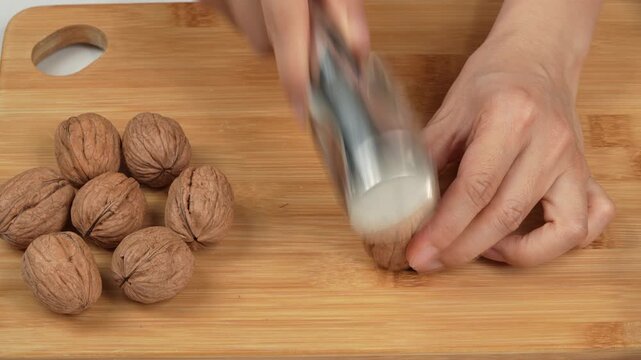 Close-up of female hands using a metal mallet to crack a whole walnut on a wooden cutting board. Authentic healthy cooking action concept.