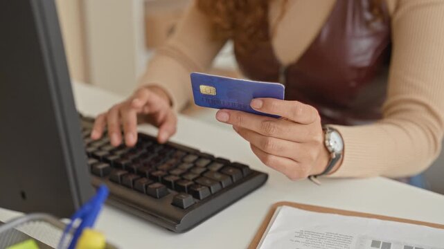Woman holding a blue creditcard with fingers while typing on a keyboard at a desk in building; online payment confidence.