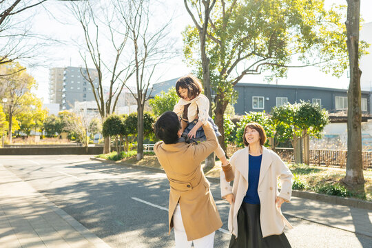 Joyful toddler girl being lifted high in the air by her parents in a winter park, happy family smiling together