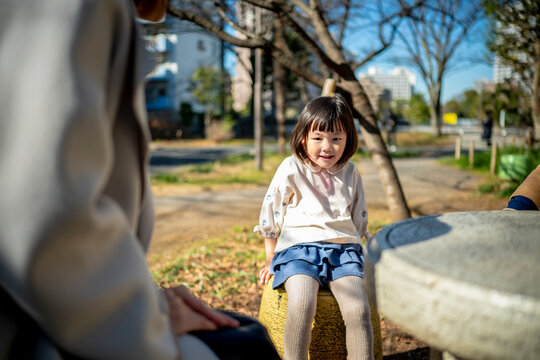 Young Japanese girl sitting on a stone stool in a park with parents in soft focus foreground