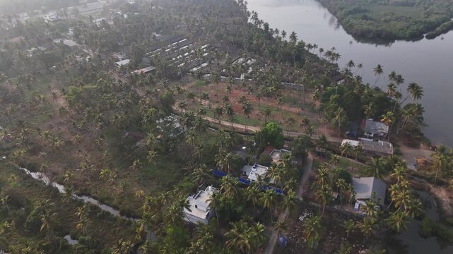 Drone footage reveals lush green coconut palm groves surrounding white residential buildings along a calm river backwater. Misty morning light illuminates the tropical landscape with red earth patches