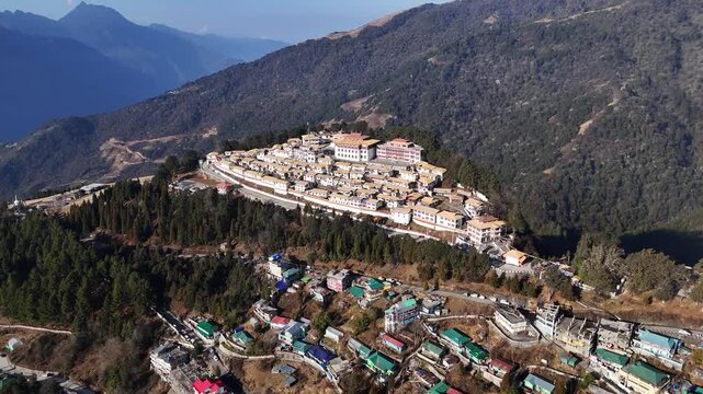 Stunning drone footage reveals the iconic Tawang Monastery complex with white buildings and golden rooftops nestled on a forested mountain ridge. Snow-capped Himalayan peaks rise in the misty blue bac