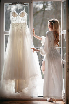 Bride in white robe looking at her wedding dress by the window
