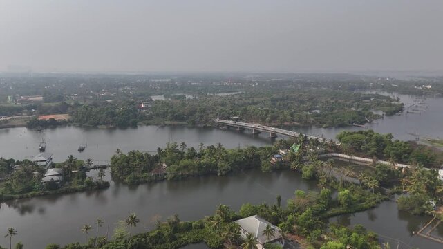 Drone footage captures the serene Kerala backwaters with a bridge crossing the river surrounded by lush green palm trees and tropical vegetation. Calm waters reflect the hazy sky as boats dock along t