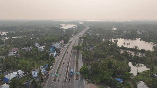 Drone footage captures a wide national highway cutting through lush green Kerala landscape dotted with coconut palm trees and serene backwaters. Vehicles travel along the multi-lane road flanked by sm