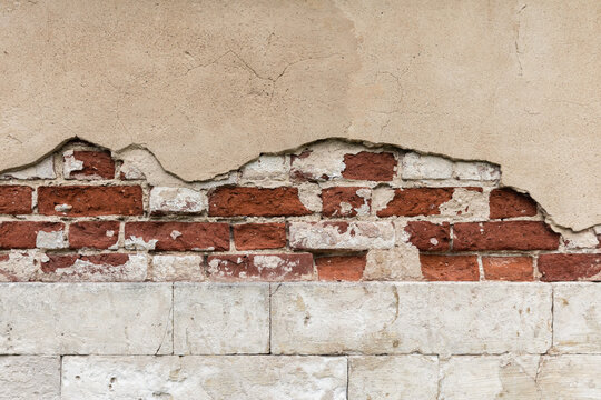 Peeling plaster reveals weathered red brick and cracked stucco on an aged urban building facade, conveying decay, texture, and rustic architectural details ideal for backgrounds and design projects