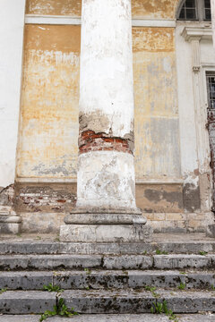 A weathered classical column stands on mossy stone steps in front of a historic building with peeling plaster, exposed brick and a crumbling facade, evoking age, decay and architectural heritage