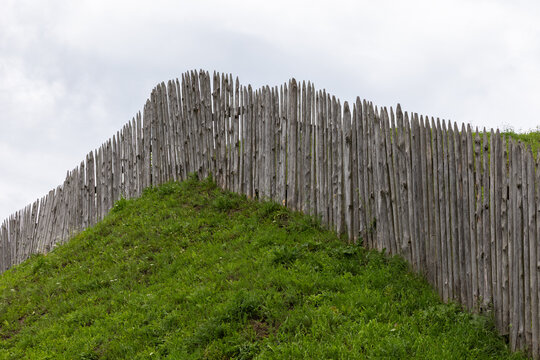 A weathered wooden palisade fence crowns a grass-covered earthwork hill beneath an overcast sky, suggesting rustic fortification, historic architecture and rural landscape textures with green grass