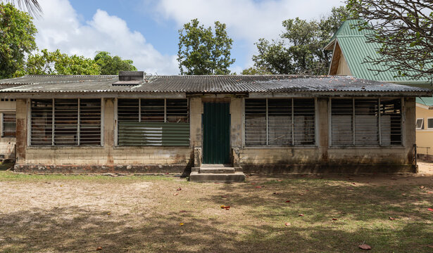 Weathered single-story school building with corrugated metal roof, louvered windows and a green door set in a sunlit tropical yard, conveying decay, history and changing community life