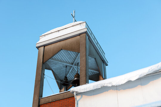 A snow-covered church bell tower with visible bells and cross set against a clear blue winter sky, conveying quiet, cold, architectural and religious atmosphere in a small town or rural setting