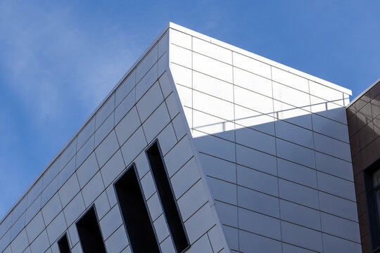 A modern white geometric building facade with clean panel lines, recessed dark windows and pronounced diagonal shadows beneath a clear blue sky, conveying minimalist architecture, contemporary design