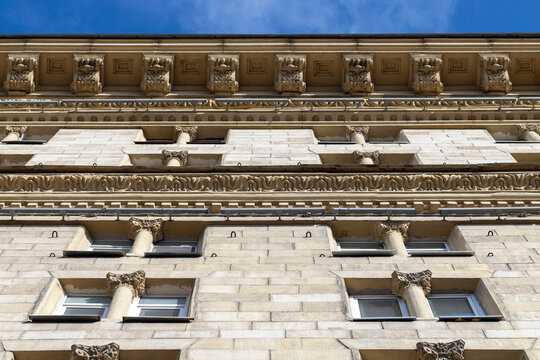 Ornate neoclassical building facade photographed looking upward, showcasing decorative columns, carved cornice and stone architectural details against a clear blue sky in an urban historic setting