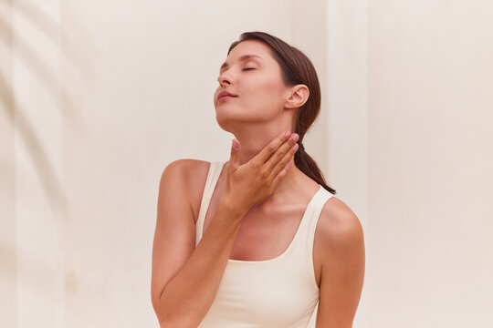 Skincare routine woman touching her neck with one hand, eyes closed and head slightly tilted in a serene expression. Clean beige column background, large copy space right. Ideal for neck firming.