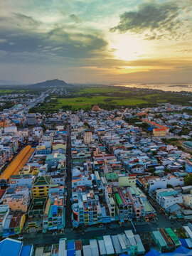 Aerial view of the dense urban landscape featuring colorful rooftops, narrow streets, and lush green rice fields under a golden sunset sky near Sam Mountain Chau Doc, An Giang, Vietnam.
