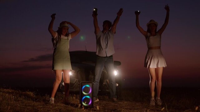 Cheerful company dances by car in evening. People friendly best close friends family on road trip stopped at night jumping partying to music next to glowing rainbow music column with flashlights.