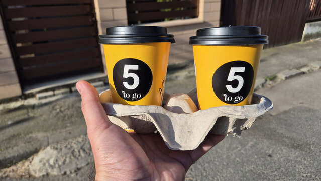 A hand holds a cardboard carrier containing two yellow "5 to go" coffee cups with black lids on a sunny day, picure taken on April 16, 2026 in Bucharest, Romania.