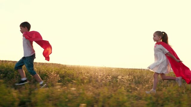 Joyful children group with red hero capes runs across rural field at back sunset light. Little kids wearing waving capes run imitating comics heroes in meadow. Cute children play superheroes in park