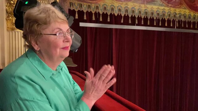 Senior happy woman is interested in watching ballet performance in theater hall, woman claps and enjoy
