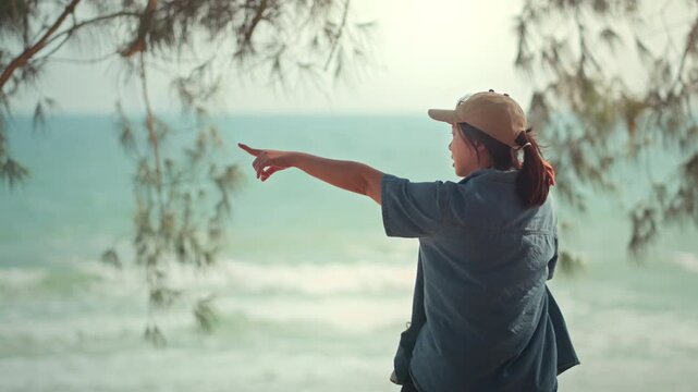 Back view of Asian mother carrying her toddler daughter and pointing at the sea. Happy family enjoying a tropical beach vacation together under the shade of trees, focusing on discovery, wellness