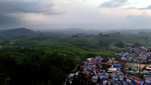 Cinematic aerial view of Valparai showcasing rolling tea-covered hills fading into misty layers, with a small town at the edge and dramatic clouds casting soft light across the landscape.