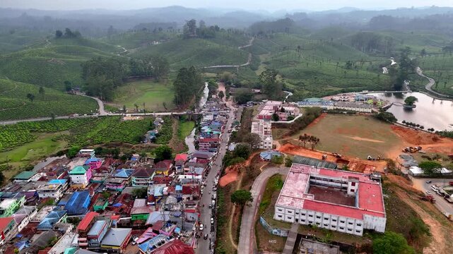 Drone footage capturing the townscape of Valparai with busy streets, mixed residential and commercial buildings, open grounds, and lush tea estates stretching into the hills.