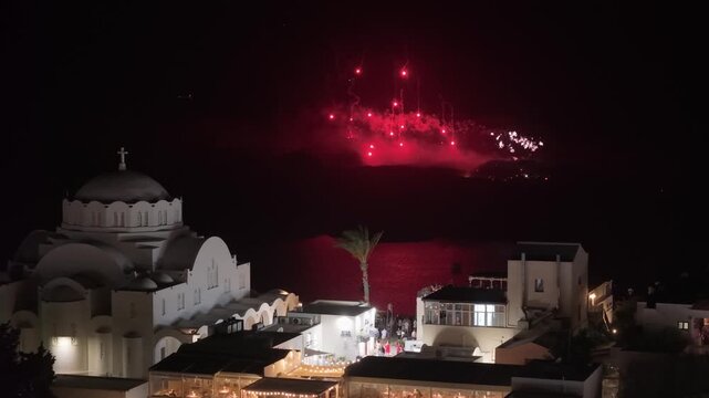 A majestic night view of the Ieros Mitropolitikos Naos Ypapantis in Thira, Santorini, with vibrant red fireworks exploding over the Nea Kameni volcano in the background.