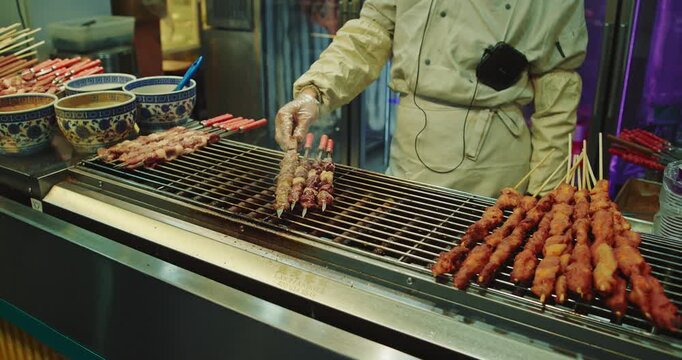 vendor in a white coat tends to rows of meat skewers sizzling on a charcoal grill at an outdoor Beijing street food market in the evening.