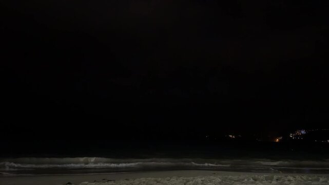 Intense lightning flash illuminating dark storm clouds over the Andaman Sea. Dramatic night scenery of Phuket beach with white ocean waves and city lights on hills, authentic monsoon photo.