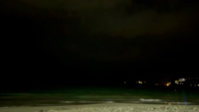 Intense lightning flash illuminating dark storm clouds over the Andaman Sea. Dramatic night scenery of Phuket beach with white ocean waves and city lights on hills, authentic monsoon photo.