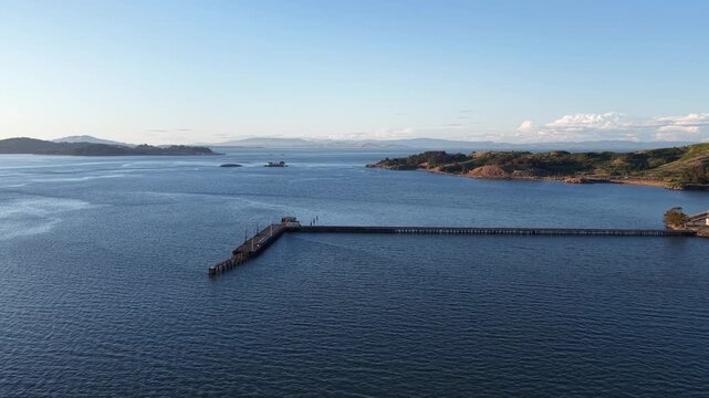 Serene aerial footage capturing a long pier stretching into the calm, blue waters of san pablo bay, with the rolling hills of the california coastline visible on the horizon under a clear sky