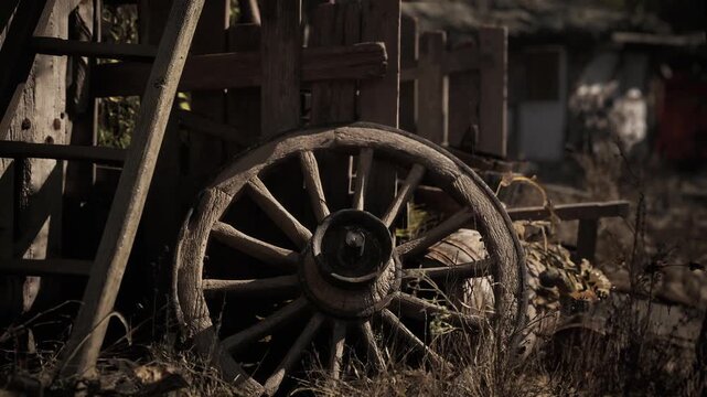 A weathered wooden wagon wheel leans against a rustic structure, surrounded by tall grass and scattered foliage, evoking a quiet atmosphere of the countryside.