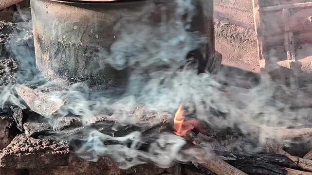 A rusty earthen chulha made of bricks sits outdoors, with thick white smoke billowing around it. Glowing firewood burns beneath a pot, heating food or water for a bath in a traditional rural way.