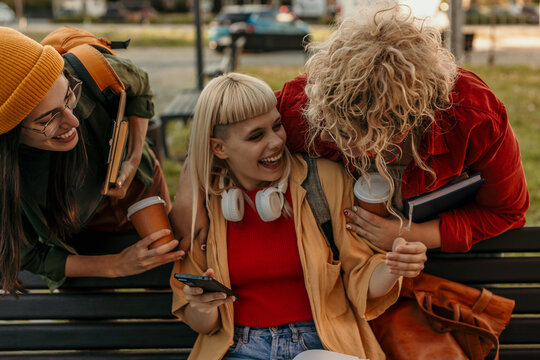 Female students laughing, enjoying coffee, and using smartphone in park