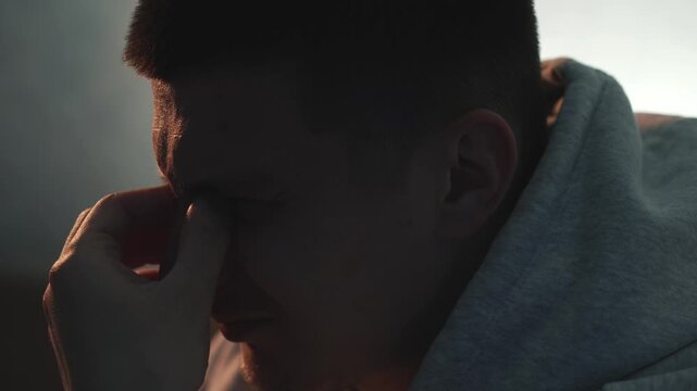 Tired man in a grey hoodie sitting in a dark room in the evening, removing his glasses and rubbing his eyes due to pain and fatigue after long computer work. Concept of eye strain, vision problems