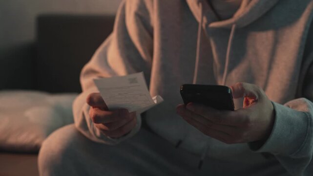 Man sitting in a dimly lit room holding a receipt and using a mobile phone for bill payment. Concept of utility bills, financial operations, banking apps, and expense management. Close-up shot of hand