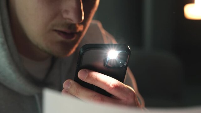 Man in grey hoodie taking photos of paper documents with a smartphone in a dim room. Concept of digital document management, scanning receipts, saving information, and modern workflow. Phone flash vis