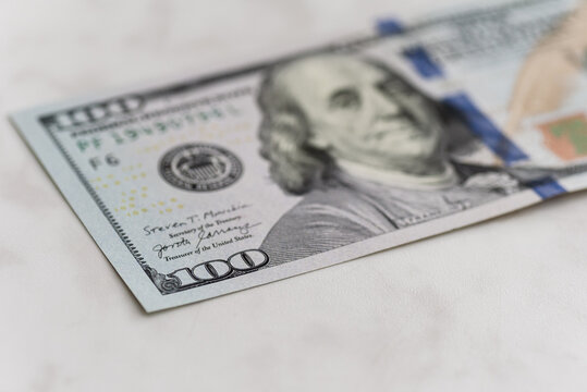 Closeup hundred dollar bill on marble surface with visible blue security ribbon, partial portrait, crisp microprinting, soft natural light, shallow depth, minimalist composition
