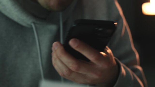 Man in a grey hoodie holding white paper documents and taking photos with a smartphone in a dark room. Concept of scanning important paperwork, digitizing files, storing receipts, and document managem