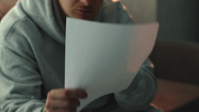 Man wearing a grey hoodie photographing paper documents with a smartphone in a dark room. He scans receipts and important papers for digital storage. Concept of everyday organization, home office 