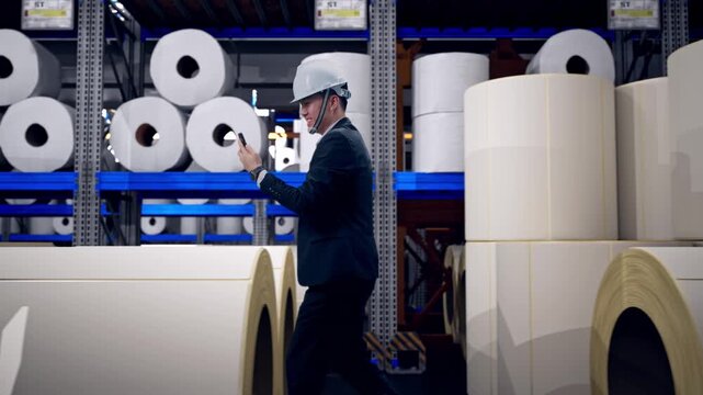 Side View of Asian Businessman Use Smartphone, Video Call While Walking at Warehouse with Rows of Stacked White Polymer and Paper Rolls