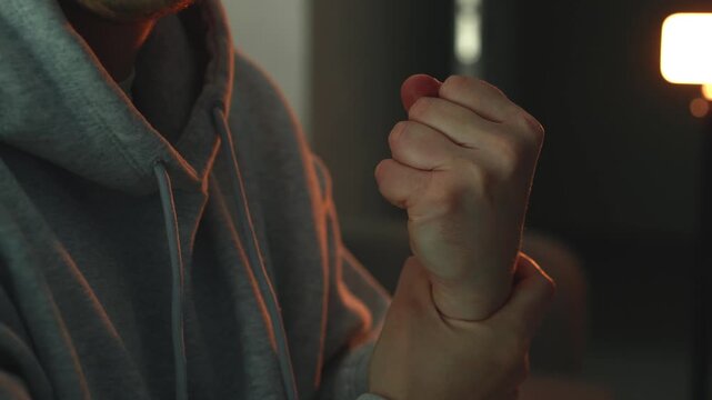 Close-up of a young man experiencing pain in his hand while sitting in a dimly lit room. He massages his wrist, makes a fist, and performs circular hand movements to relieve discomfort.