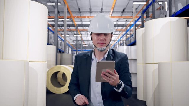 Front View of Asian Businessman Use Tablet While Walking at Warehouse with Rows of Stacked White Polymer and Paper Rolls