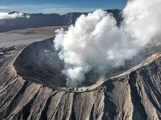 Fototapeta premium Aerial view of the Bromo volcano's caldera emitting a plume of white smoke, a stark contrast against the dark, rugged terrain, Mt. Bromo, East Java, Indonesia