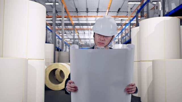 Front View of Asian Businessman Looking At Blueprint In His Hands While Walking at Warehouse with Rows of Stacked White Polymer and Paper Rolls