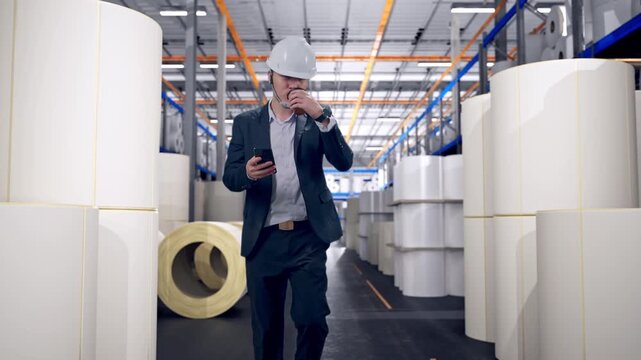 Front View of Asian Businessman Use Smartphone, Drink Coffee While Walking at Warehouse with Rows of Stacked White Polymer and Paper Rolls