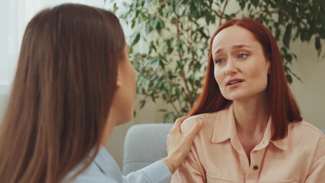 Emotional woman wiping tears with tissue while talking to female psychologist during therapy session in calm office environment with support
