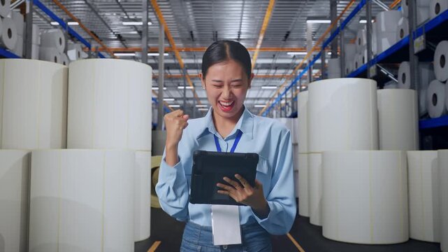 Asian Female With Her Tablet at Warehouse with Rows of Stacked White Polymer and Paper Rolls, She Raises Her Fist Up With Screaming Goal