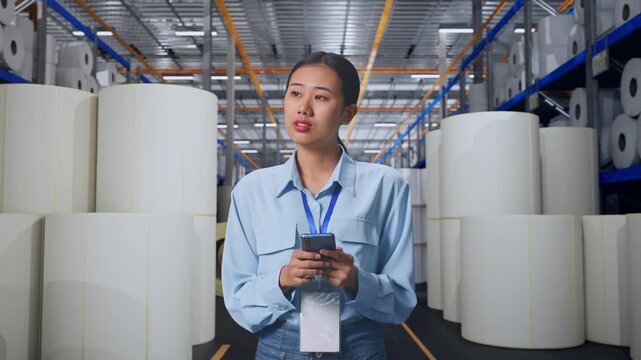 Asian Female With Her Smarphone at Warehouse with Rows of Stacked White Polymer and Paper Rolls, She Observes By Looking Around Before She Come To Concentrating With Her Smarphone